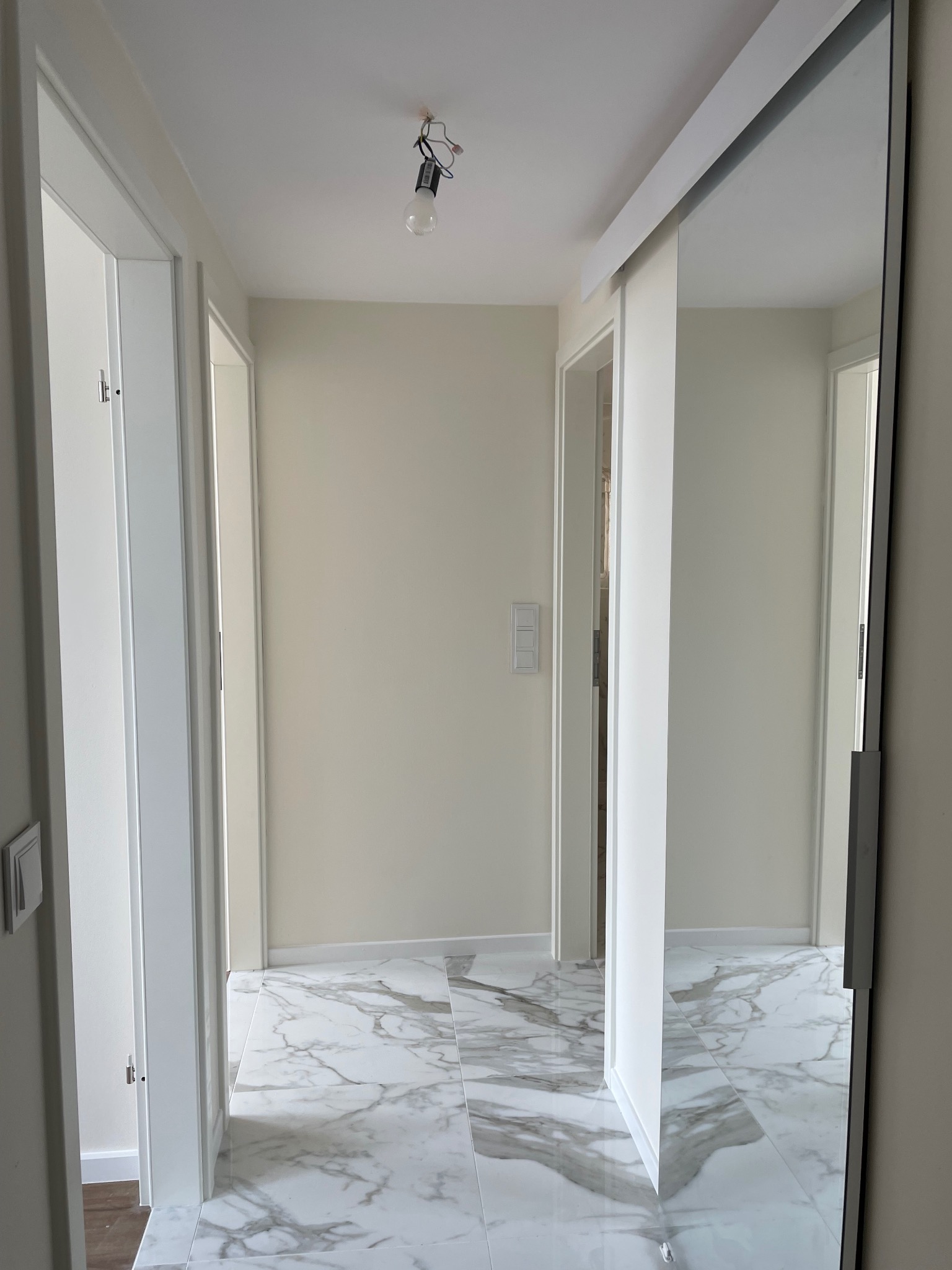 Wnętrze korytarza z białymi ścianami, marmurową podłogą i dużym przesuwnym lustrem; widoczna surowa żarówka zwisająca z sufitu.