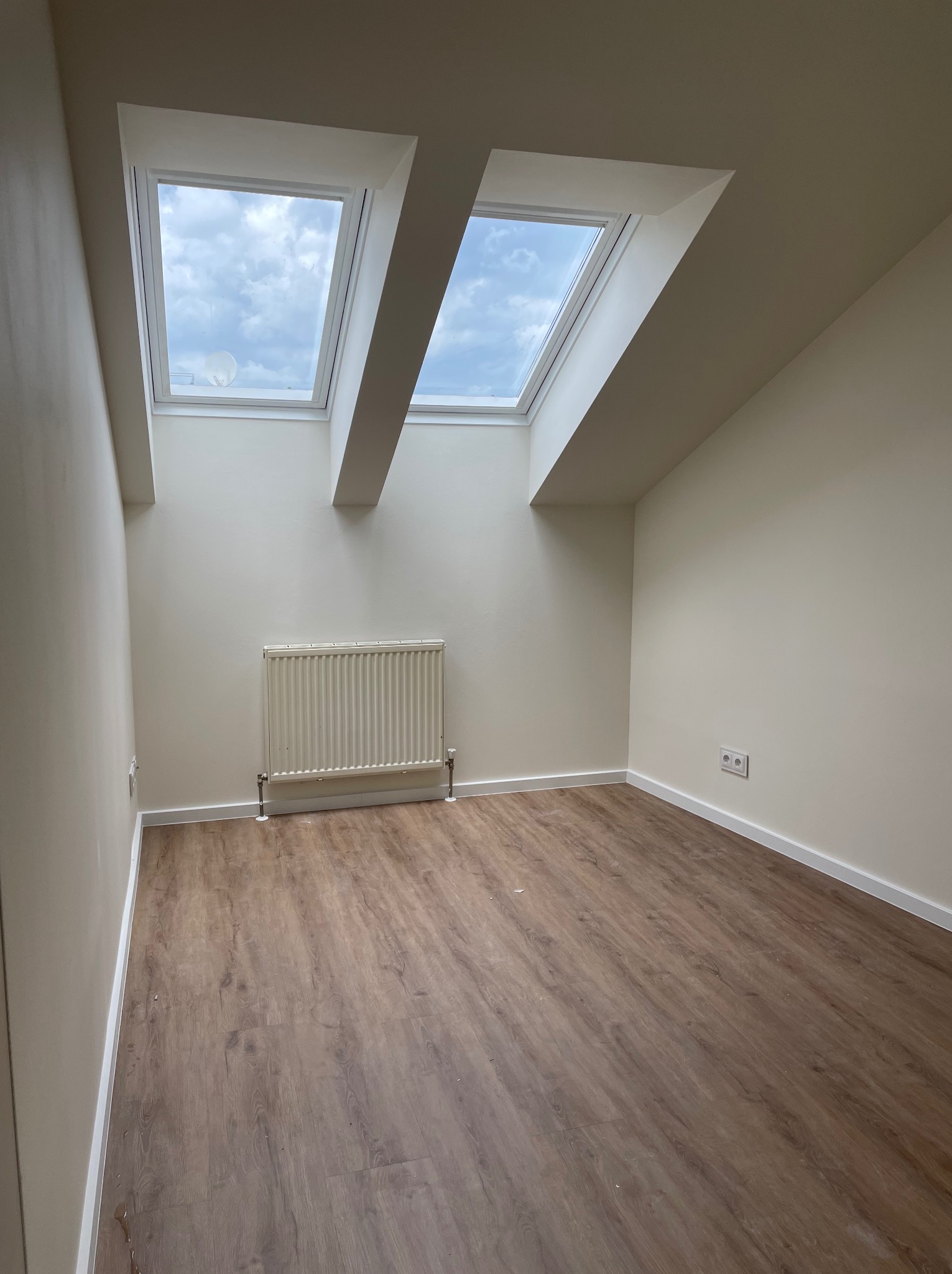 Wnętrze pokoju na poddaszu z drewnopodobną podłogą, świeżo pomalowanymi ścianami w odcieniach beżu, grzejnikiem pod dwoma oknami dachowymi i gniazdkiem elektrycznym na ścianie.