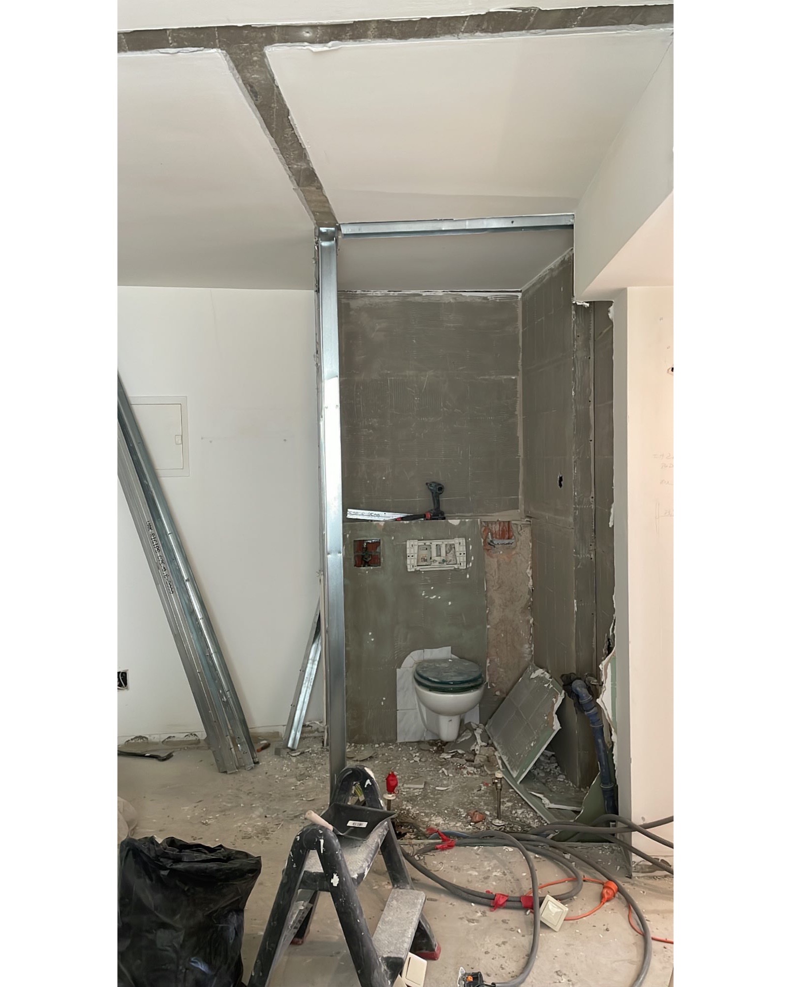 Wnętrze łazienki w trakcie prac remontowych: widoczne metalowe profile konstrukcyjne, częściowo położone szare płytki, wisząca muszla klozetowa, przewody elektryczne, narzędzia i gruz na podłodze.