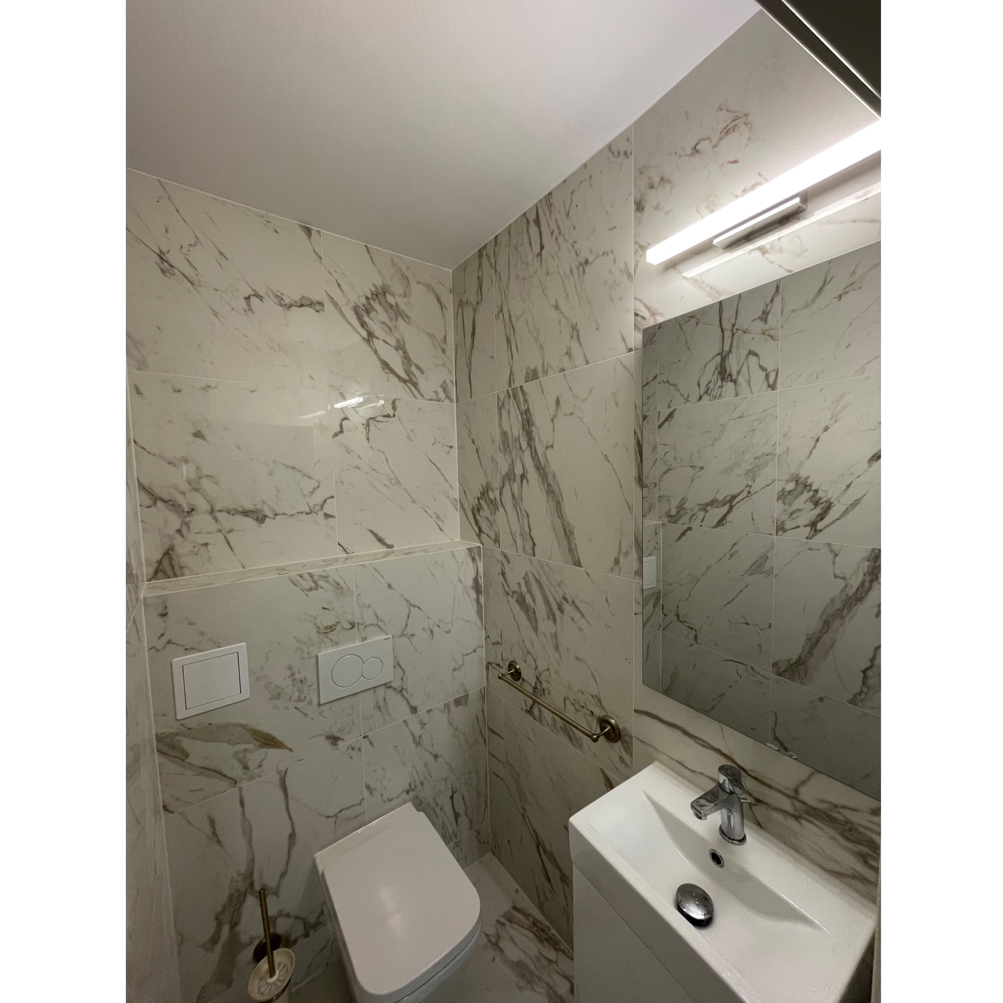 Nowoczesna łazienka wykończona płytkami imitującymi marmur, z podwieszaną toaletą, umywalką nablatową i chromowaną armaturą. Na ścianie widoczny włącznik i uchwyt na ręcznik w kolorze starego złota.