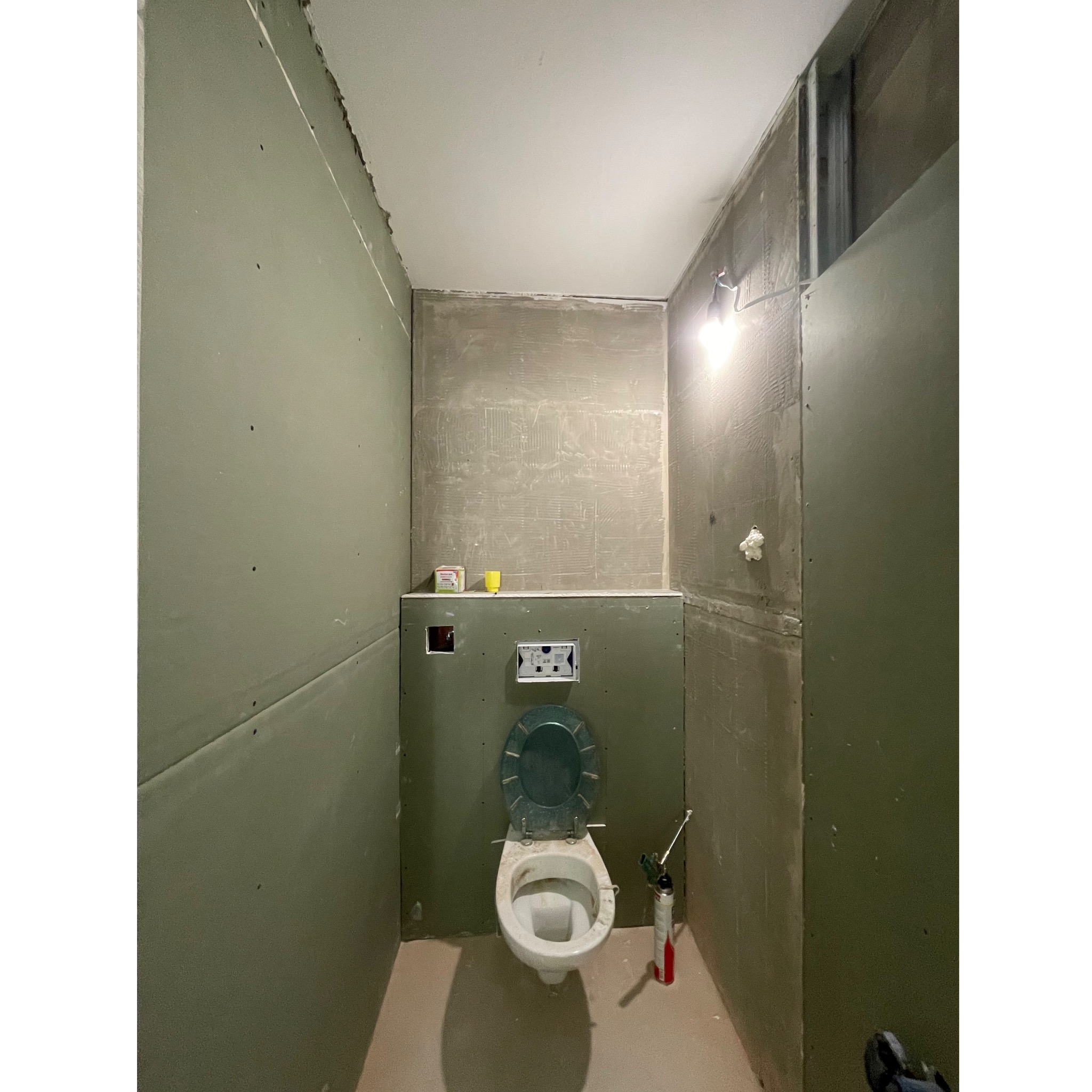 Łazienka w trakcie remontu: widoczne płyty gipsowo-kartonowe na ścianach, przygotowane do dalszych prac wykończeniowych, oraz częściowo zamontowana, brudna toaleta z otwartą klapą i pistolet...