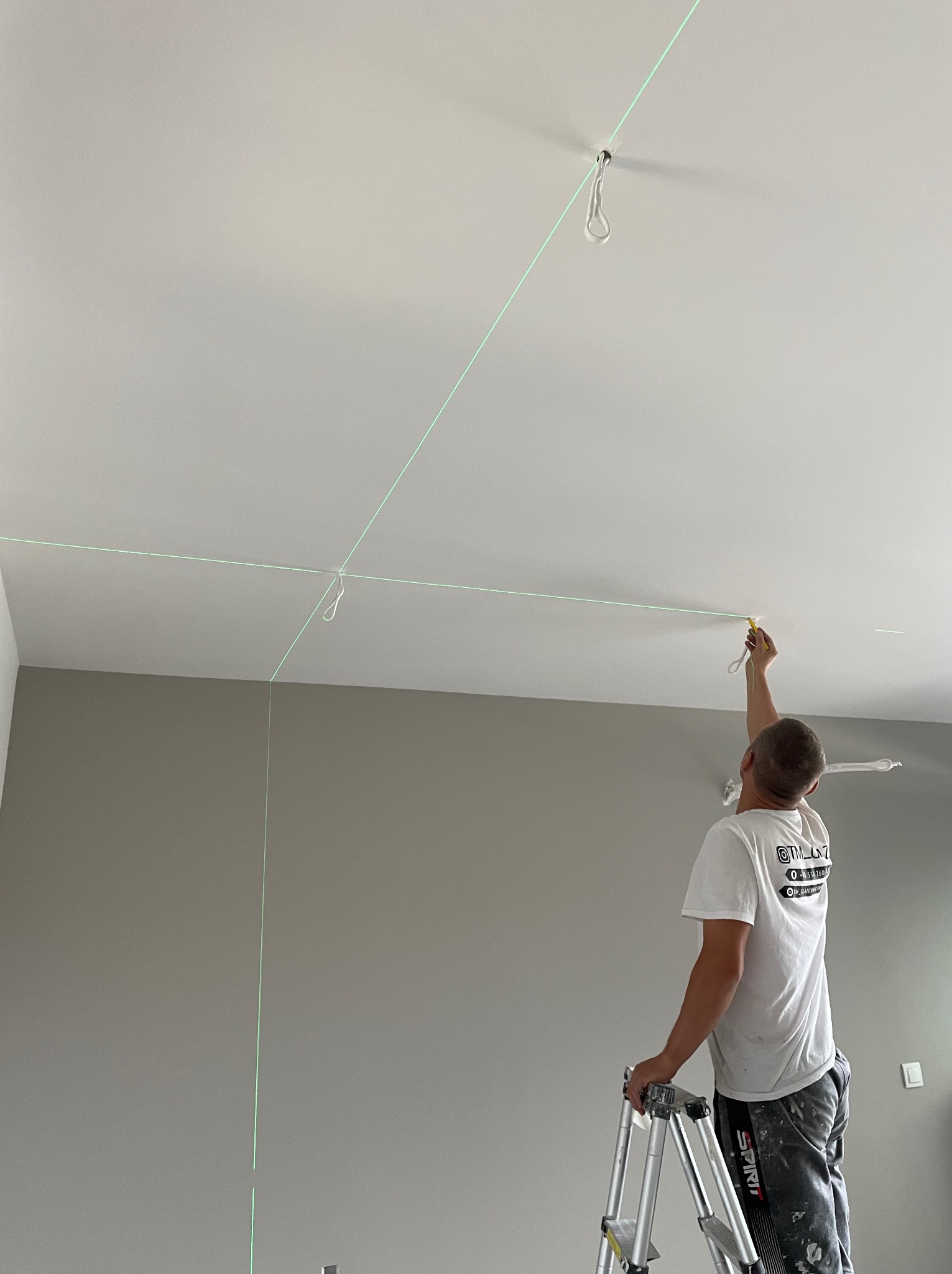 Pracownik na drabinie, wyznaczający laserowo poziom na suficie w szarym pomieszczeniu, przygotowującym do dalszych prac wykończeniowych.