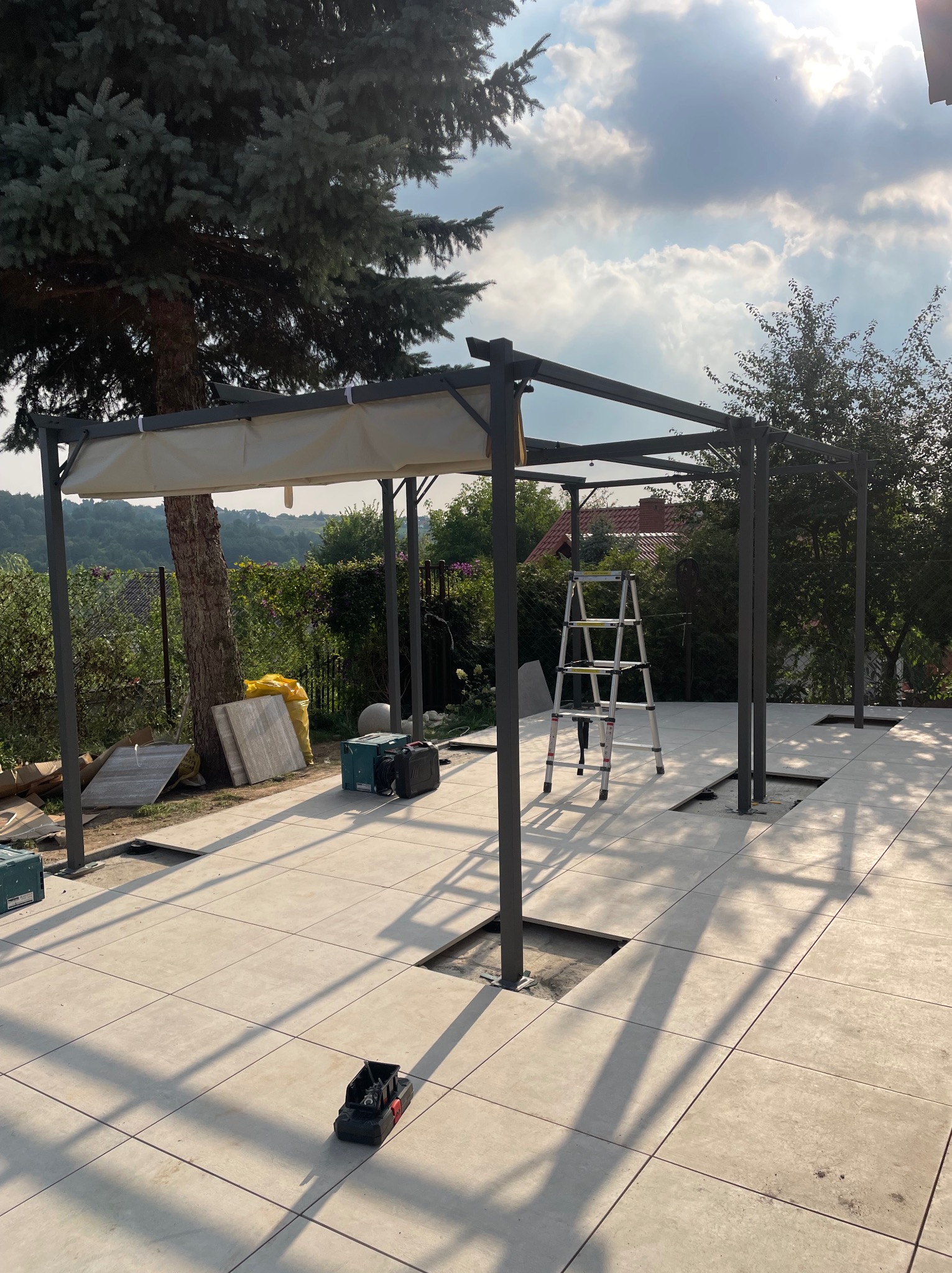 Montaż metalowej konstrukcji z zadaszeniem na tarasie w Krakowie, widoczne płytki tarasowe, drabina i narzędzia, w tle zieleń ogrodu i błękitne niebo.