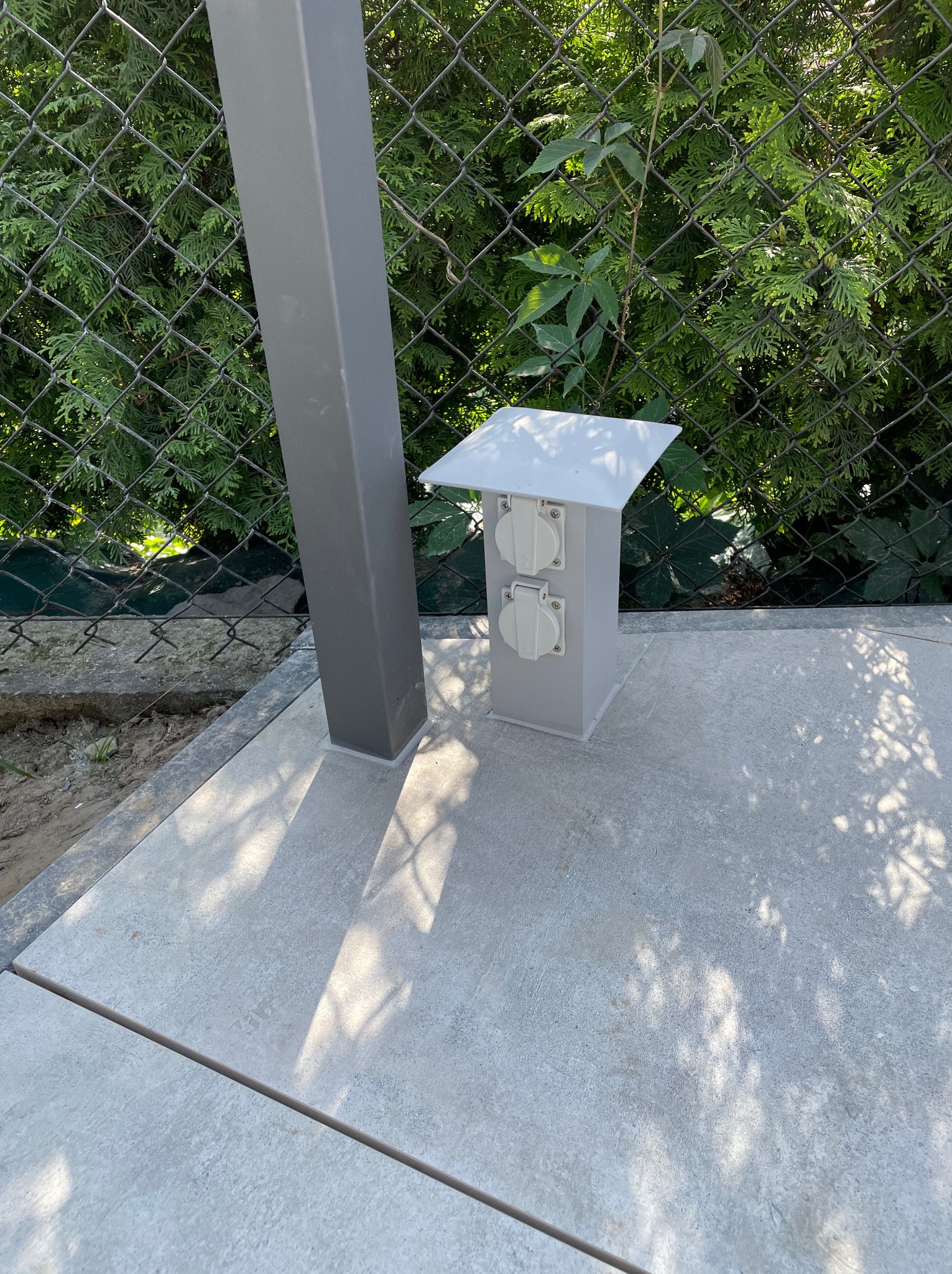 Zewnętrzna, podwójna skrzynka z gniazdami elektrycznymi, z daszkiem ochronnym, zamontowana na płytach tarasowych przy ogrodzeniu z siatki.