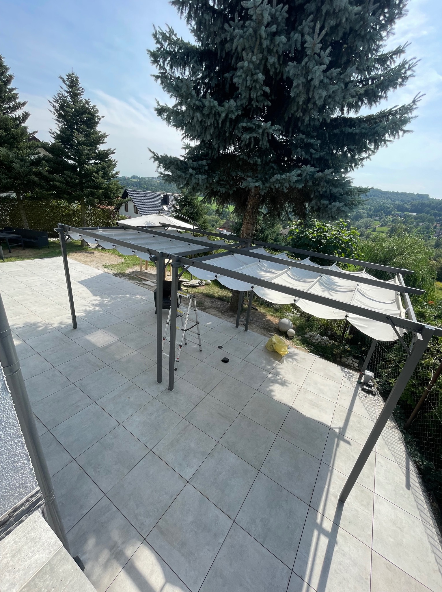 Montaż pergoli z rozsuwanym zadaszeniem na tarasie wyłożonym szarą, kwadratową płytką; widoczna drabina i narzędzia, w tle drzewa i zabudowa jednorodzinna.