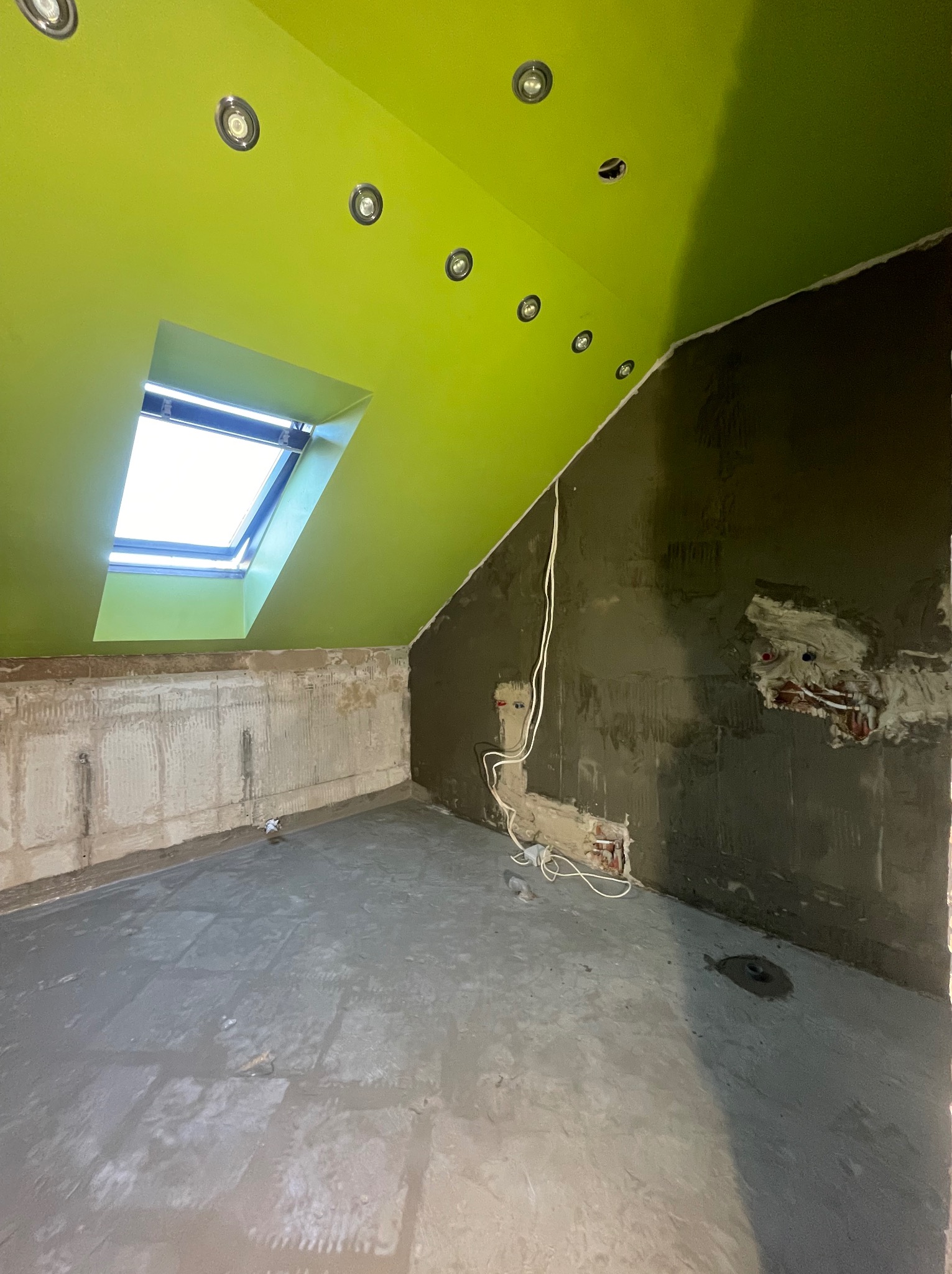 Pokój na poddaszu w trakcie prac wykończeniowych: świeżo wylana wylewka na podłodze, fragment ściany z nałożoną szarą zaprawą, widoczne przewody elektryczne wystające ze ściany, zielony sufit...