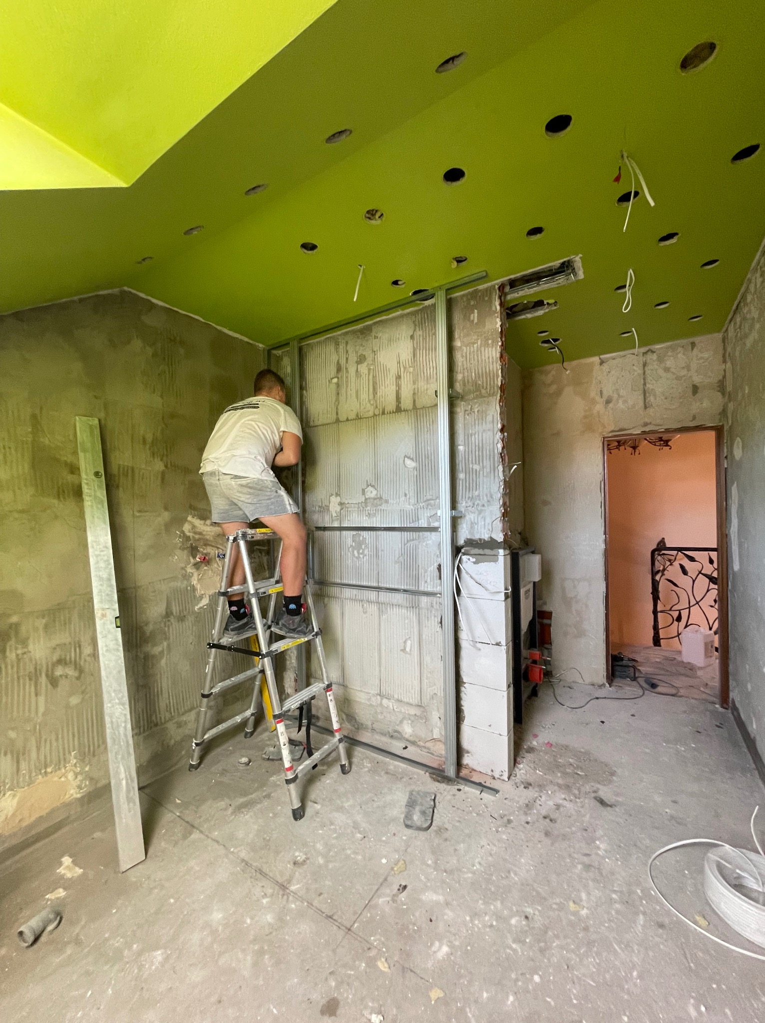 Mężczyzna na drabinie montuje metalową konstrukcję na ścianie w trakcie remontu pomieszczenia z zielonym sufitem i widocznymi przewodami elektrycznymi.
