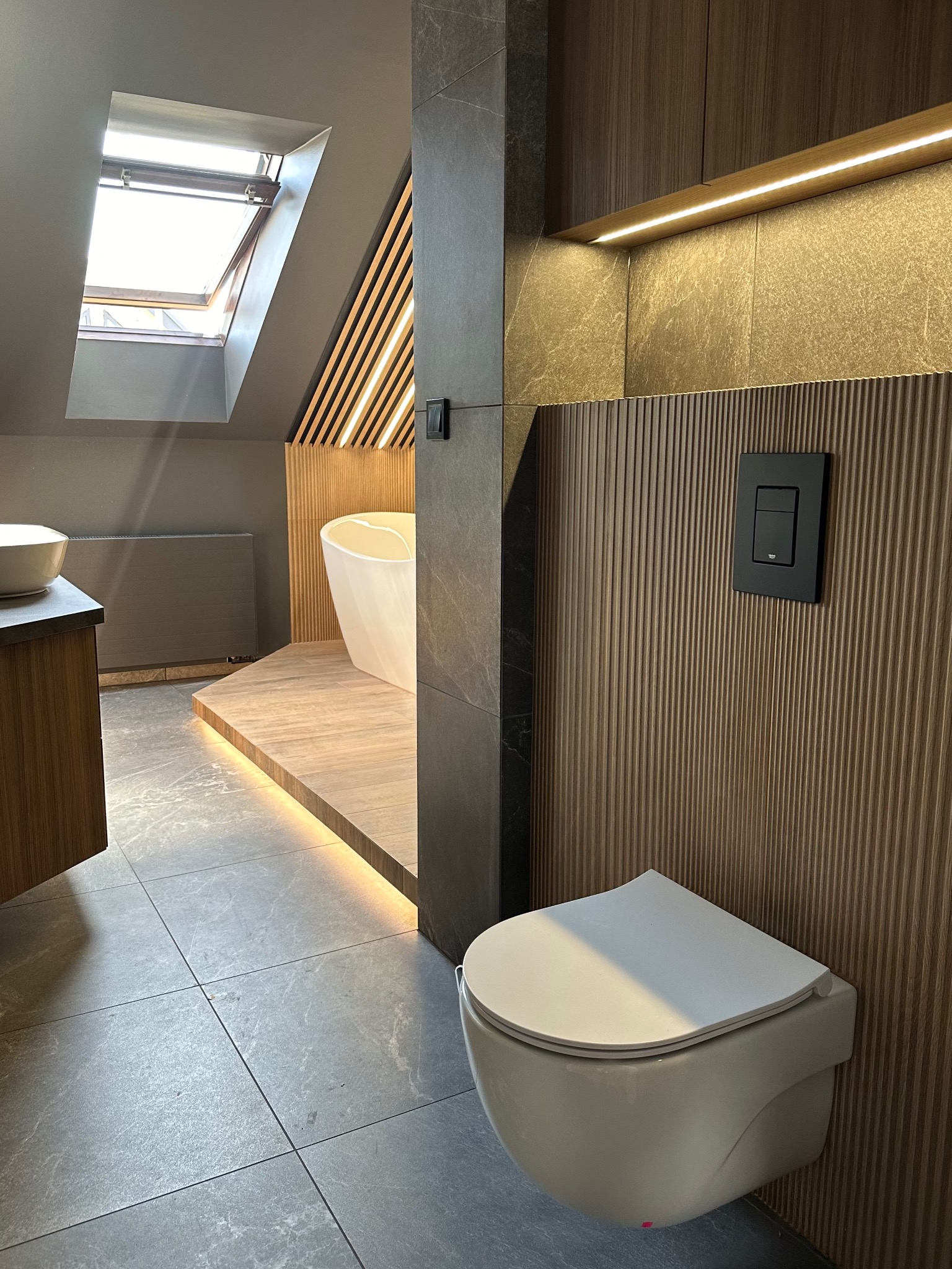 Nowoczesna łazienka na poddaszu z wolnostojącą wanną, podświetlanym podestem, wiszącą toaletą i szarą płytką podłogową