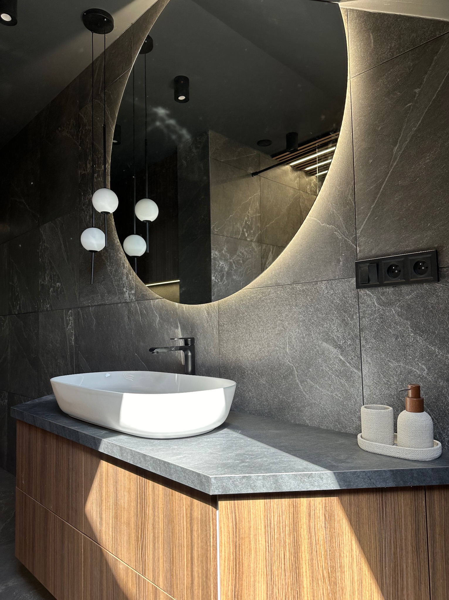 Nowoczesna łazienka z umywalką nablatową, podświetlanym lustrem owalnym i ciemnymi płytkami imitującymi kamień; widoczne wiszące lampy kuliste i drewnopodobna szafka pod umywalką.