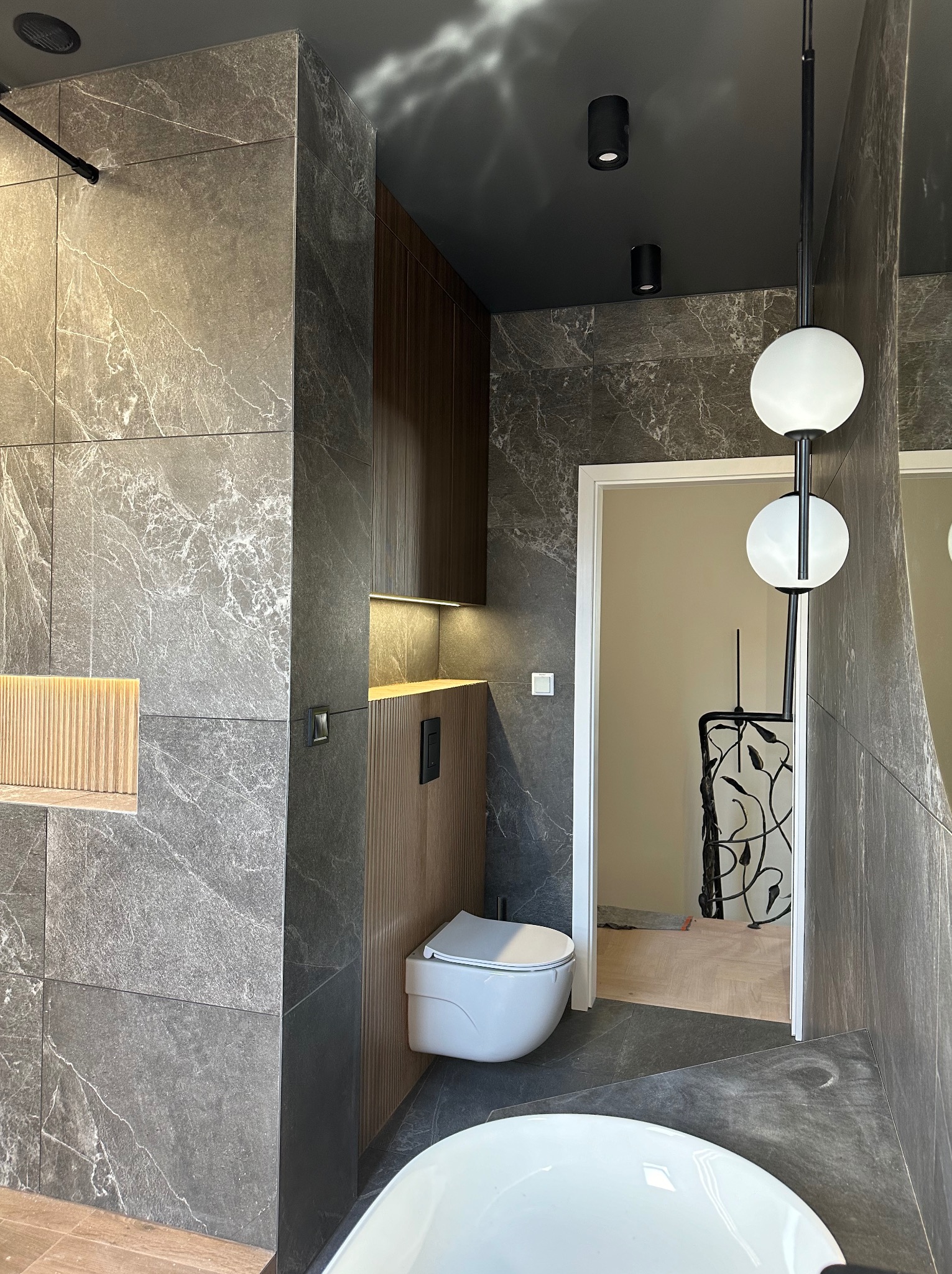 Nowoczesna łazienka z szarymi płytkami imitującymi marmur, podwieszaną toaletą z drewnianą obudową, oświetleniem LED i dekoracyjną lampą kulistą.