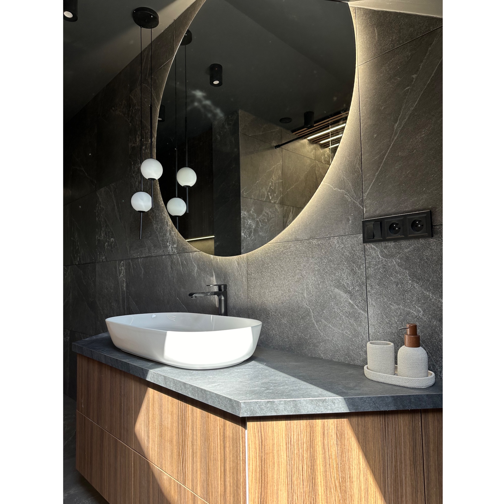 Nowoczesna łazienka z owalnym lustrem z podświetleniem LED, białą umywalką nablatową, armaturą w kolorze czarnym oraz szarymi płytkami imitującymi kamień na ścianach.