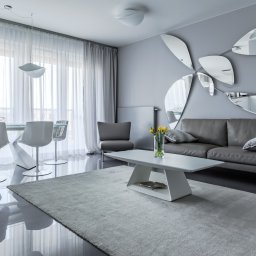 Interio design - Nowoczesny salon w odcieniach szarości z lustrzaną dekoracją ścienną w kształcie kwiatu, białym dywanem i designerskimi krzesłami.