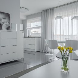 Interio design - Jasne, minimalistyczne wnętrze: białe meble, szklany stół z krzesłami, żółte narcyzy w wazonie na pierwszym planie, na komodzie czarno-białe zdjęcie dziecka i świeczniki.