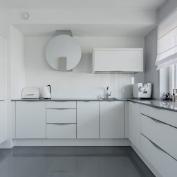 Interio design - Minimalistyczna, biała kuchnia z szarymi blatami i połyskującą, szarą podłogą; widoczne zabudowane piekarniki, okrągły okap i ekspres do kawy przy oknie z roletą rzymską.