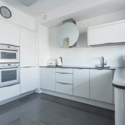 Interio design - Biała, nowoczesna kuchnia z szarymi płytkami podłogowymi i blatem, wyposażona w sprzęt AGD: piekarnik, mikrofalówkę, ekspres do kawy oraz okrągły okap.