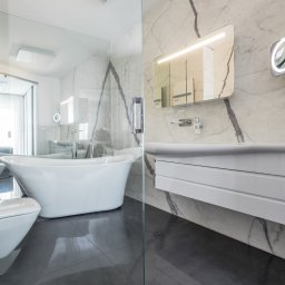 Interio design - Nowoczesna łazienka z marmurowymi ścianami, wolnostojącą wanną, podwieszaną toaletą i szafką z umywalką, oświetlona lustrami.