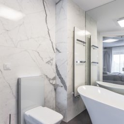 Interio design - Nowoczesna, minimalistyczna łazienka z białymi marmurowymi ścianami, podwieszaną toaletą, grzejnikiem drabinkowym i wolnostojącą wanną, odbijającą się w lustrze.