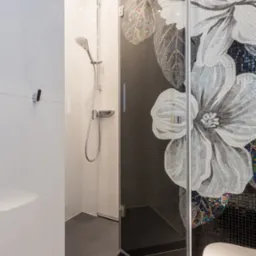 Nowoczesna kabina prysznicowa z czarną mozaiką i motywem kwiatowym z jasnych płytek, chromowana armatura, białe płytki na ścianach, szara podłoga.