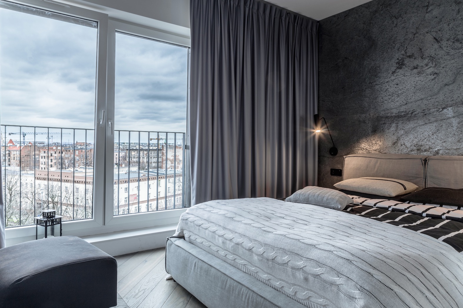 Nowoczesna sypialnia z łóżkiem w odcieniach szarości, dekoracyjną ścianą imitującą kamień i widokiem na miasto z okna balkonowego.
