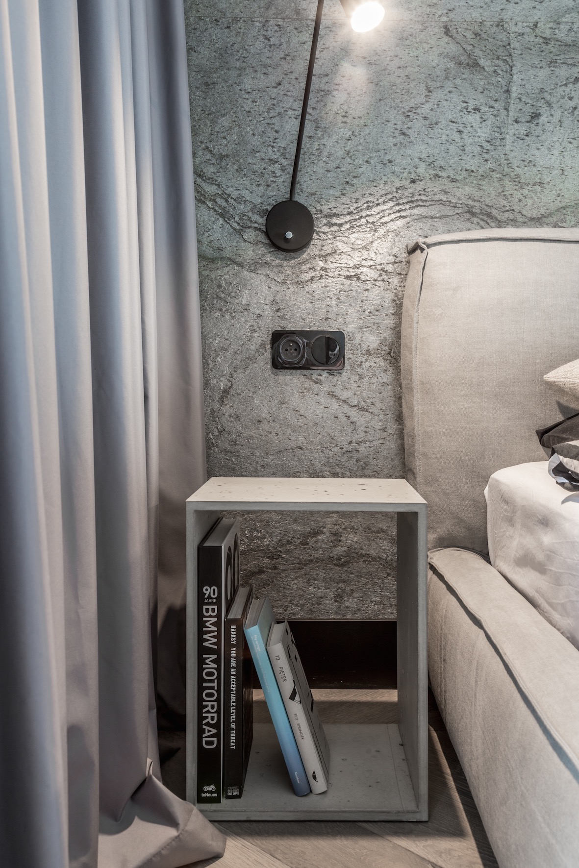 Szary pokój z łóżkiem, betonową ścianą z lampą ścienną i gniazdkiem elektrycznym, oraz szafką nocną z książkami: BMW Motorrad, Banksy, 13 Pięter.