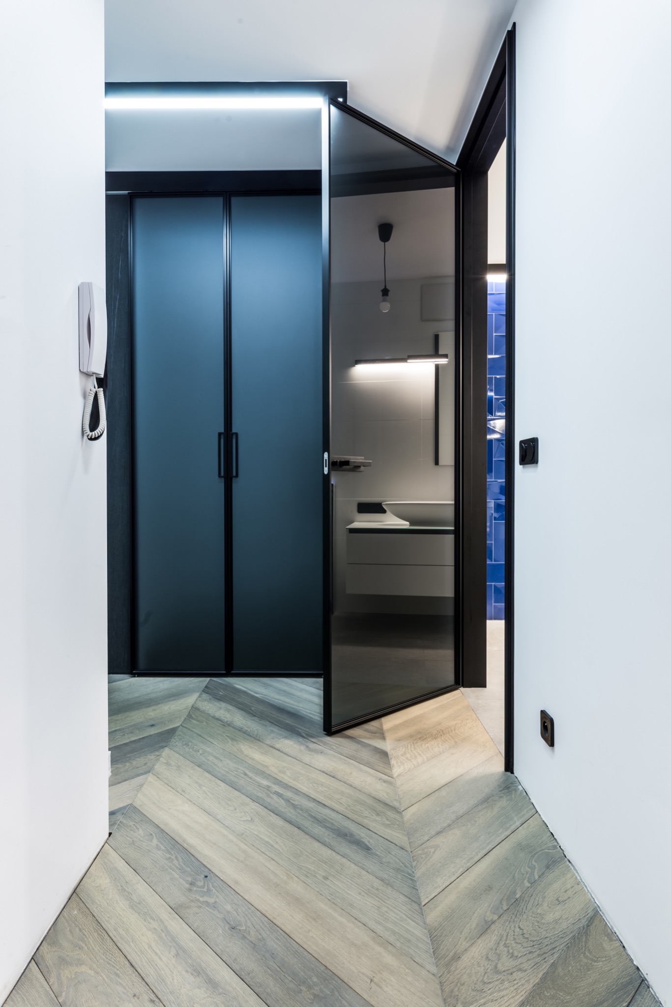 Nowoczesny przedpokój z parkietem w jodełkę, prowadzący do łazienki z niebieskimi płytkami, widocznej przez szklane drzwi.