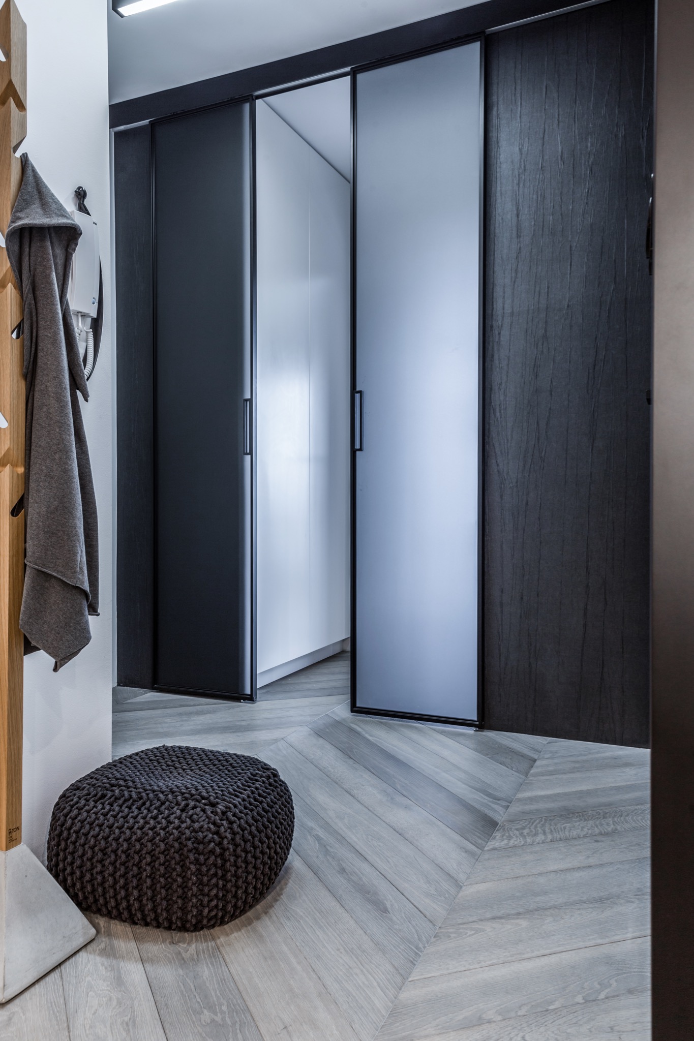 Nowoczesny przedpokój z szarymi, przesuwnymi drzwiami, pufą z dzianiny i drewnianym wieszakiem z szarym szlafrokiem na tle jasnej podłogi ułożonej w jodełkę.