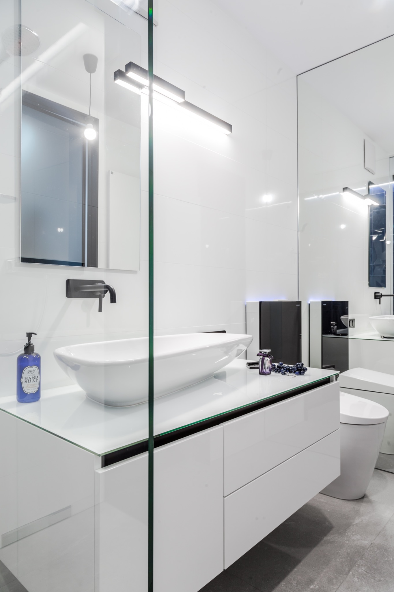 Nowoczesna, biała łazienka z umywalką nablatową, czarną armaturą, szklaną kabiną prysznicową i dekoracyjnymi akcentami w postaci ciemnych koralików i flakonika perfum na blacie.