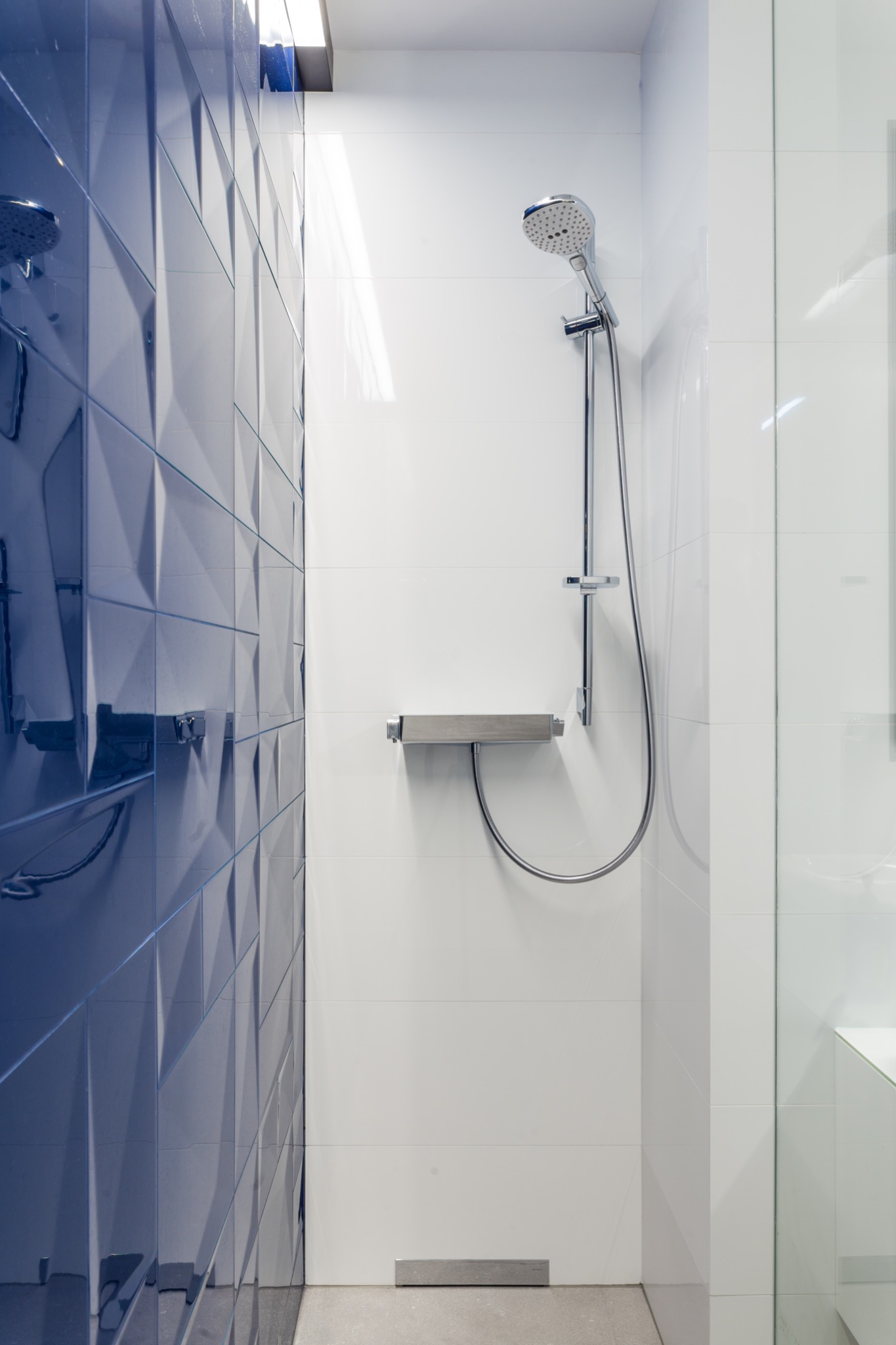 Nowoczesna kabina prysznicowa z niebieskimi, trójwymiarowymi płytkami na jednej ścianie i białymi płytkami na pozostałych, chromowana armatura, odpływ liniowy w posadzce.