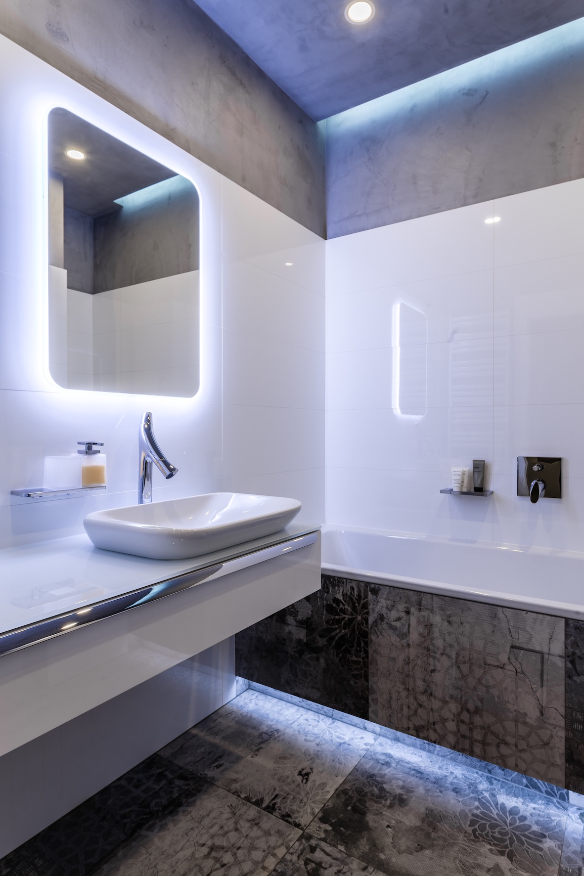 Nowoczesna łazienka z podświetlanym lustrem, białą umywalką nablatową i szarymi płytkami z motywem kwiatowym podświetlonymi od dołu.