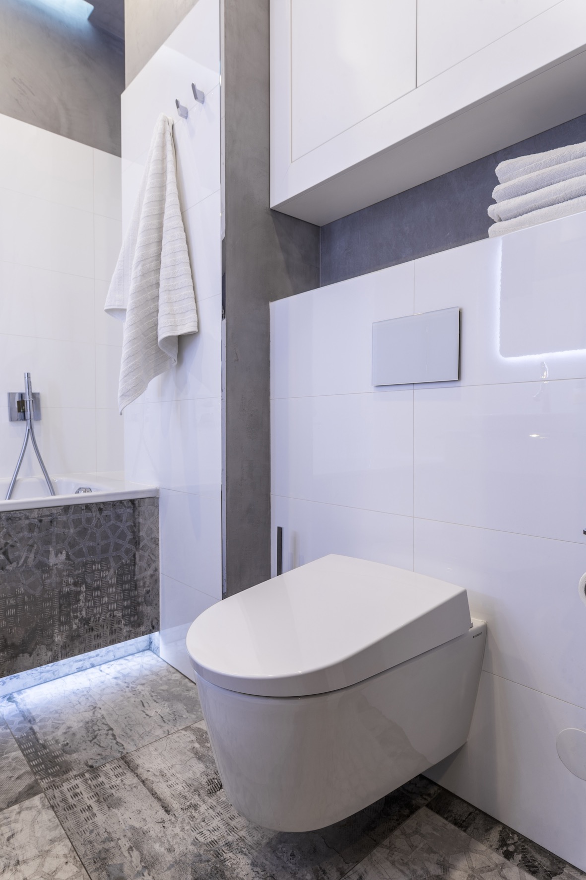 Nowoczesna łazienka w bieli i szarościach z podwieszaną toaletą, płytkami imitującymi beton na podłodze i podświetleniem LED wanny.