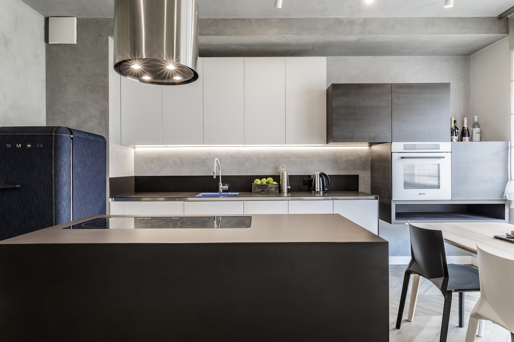 Nowoczesna kuchnia w odcieniach szarości z wyspą kuchenną, białymi szafkami, sprzętem AGD marki SMEG, w tym lodówką obitą materiałem imitującym denim, oraz oświetleniem LED pod szafkami.