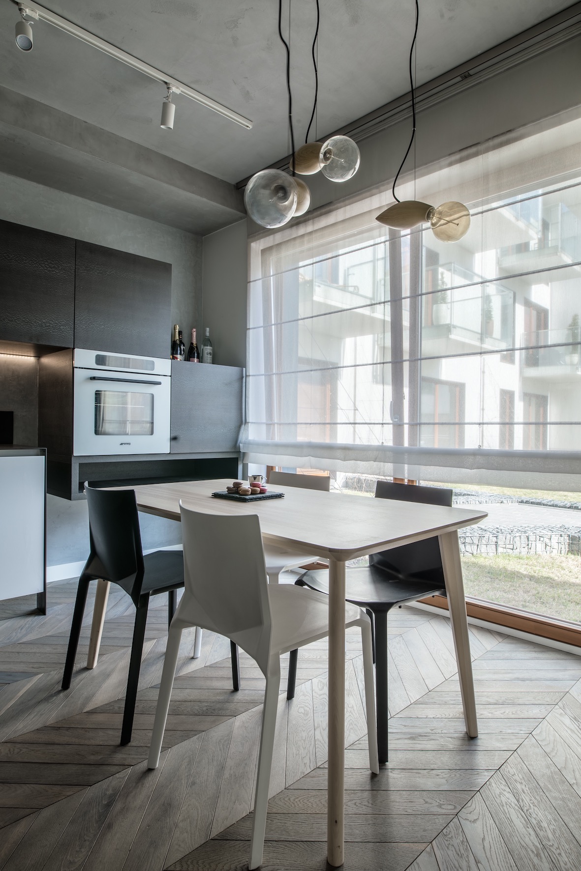 Nowoczesna jadalnia z drewnianym stołem, czarnymi i białymi krzesłami, parkietem ułożonym w jodełkę, oświetleniem punktowym i wiszącymi lampami, widokiem na budynek przez okno z roletami rzymskimi.