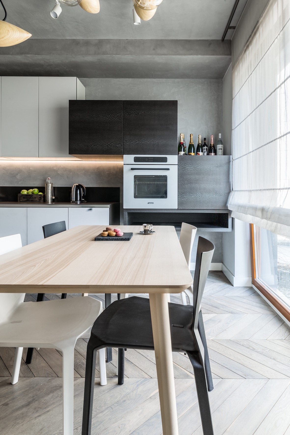 Nowoczesna kuchnia z drewnianym stołem, czarnymi i białymi krzesłami, oraz minimalistycznymi szafkami w odcieniach szarości i czerni, oświetlona designerskimi lampami sufitowymi.