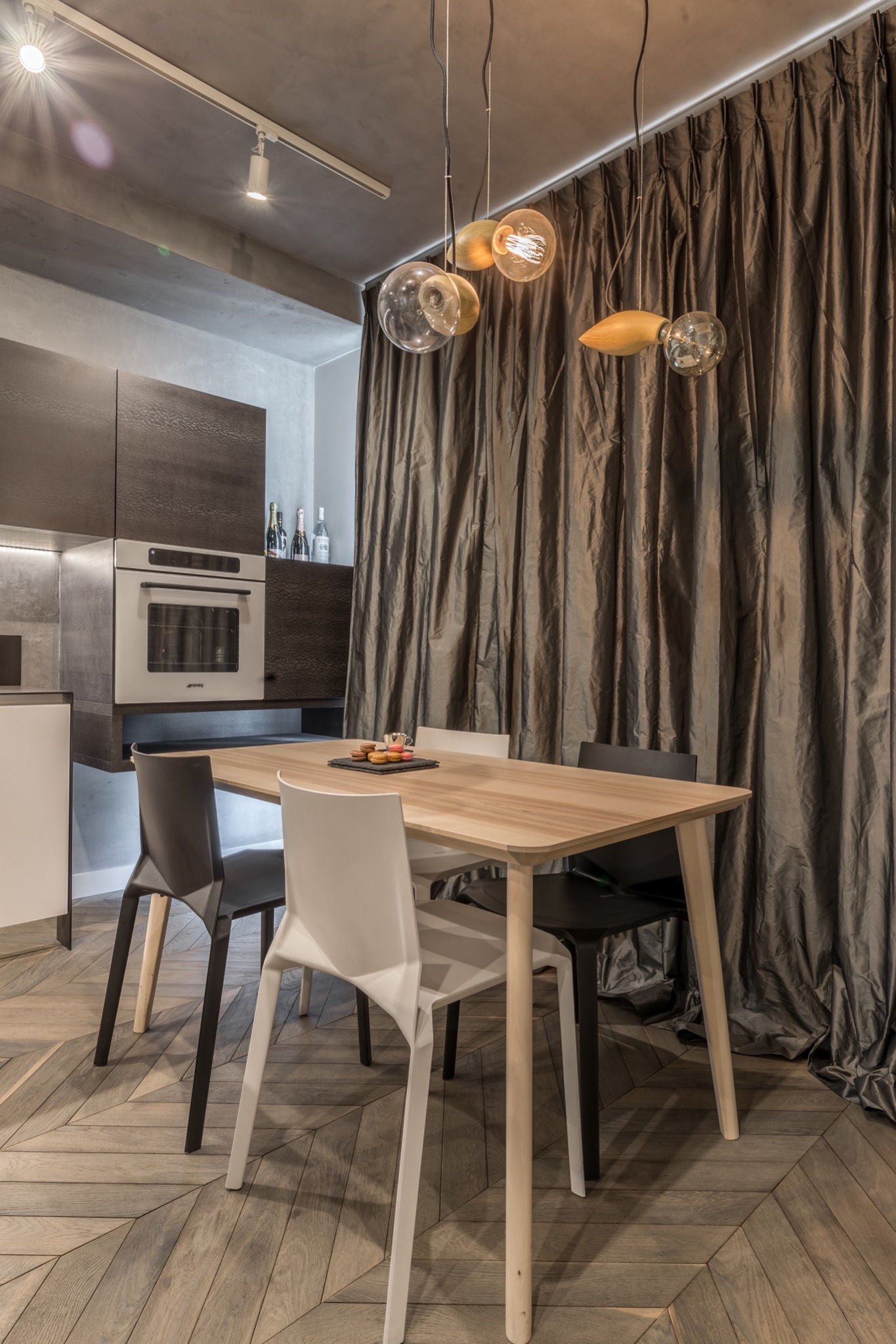 Jadalnia z drewnianym stołem, czarnymi i białymi krzesłami, parkietem w jodełkę i dekoracyjnymi lampami wiszącymi na tle drapowanej, błyszczącej zasłony.