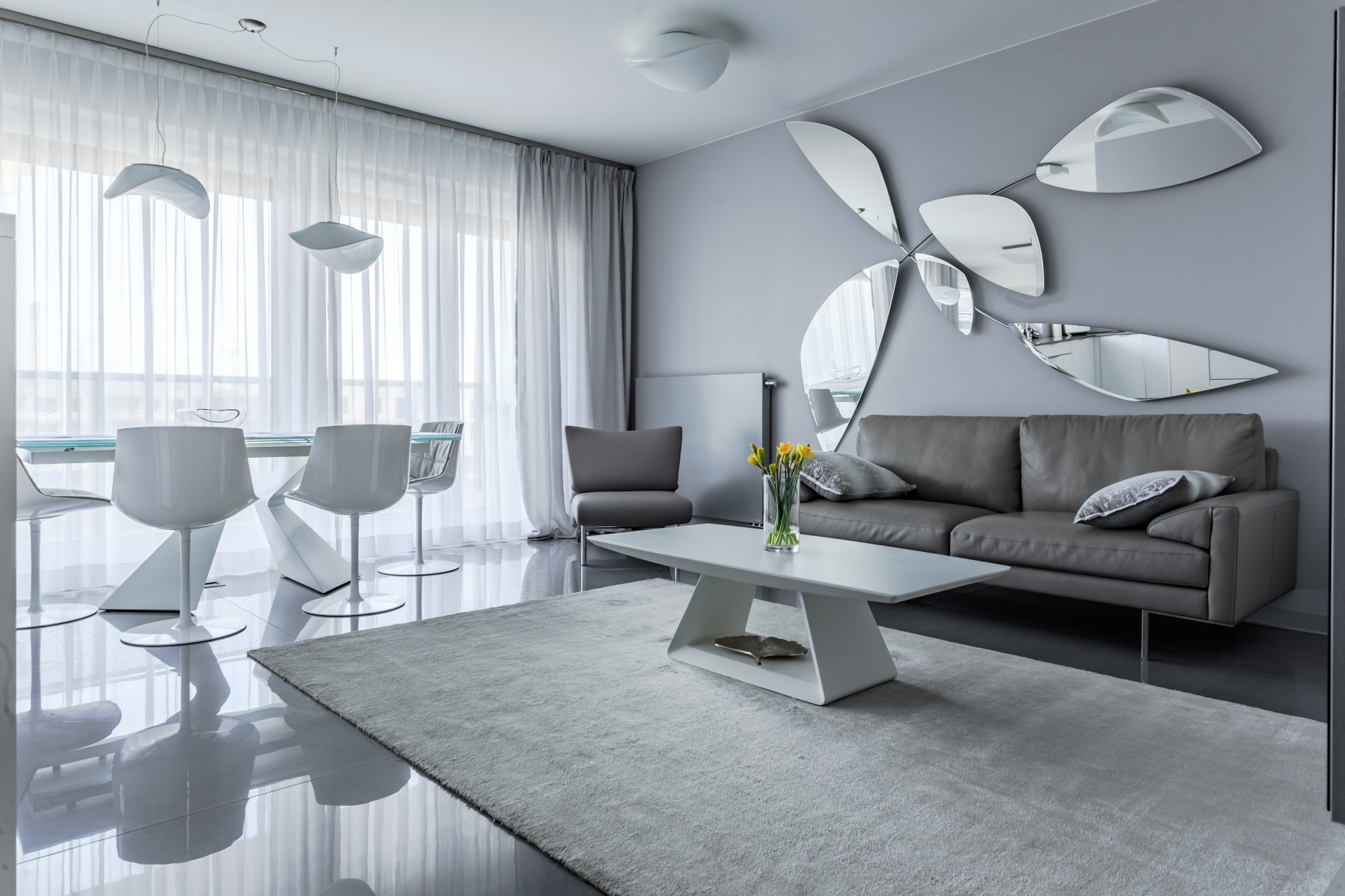 Nowoczesny salon w odcieniach szarości z lustrzaną dekoracją ścienną w kształcie kwiatu, białym dywanem i designerskimi krzesłami.