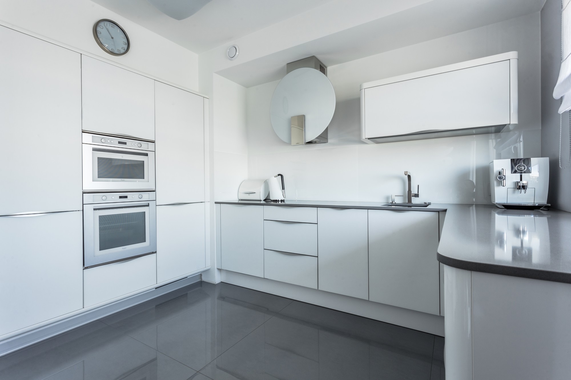 Biała, nowoczesna kuchnia z szarymi płytkami podłogowymi i blatem, wyposażona w sprzęt AGD: piekarnik, mikrofalówkę, ekspres do kawy oraz okrągły okap.