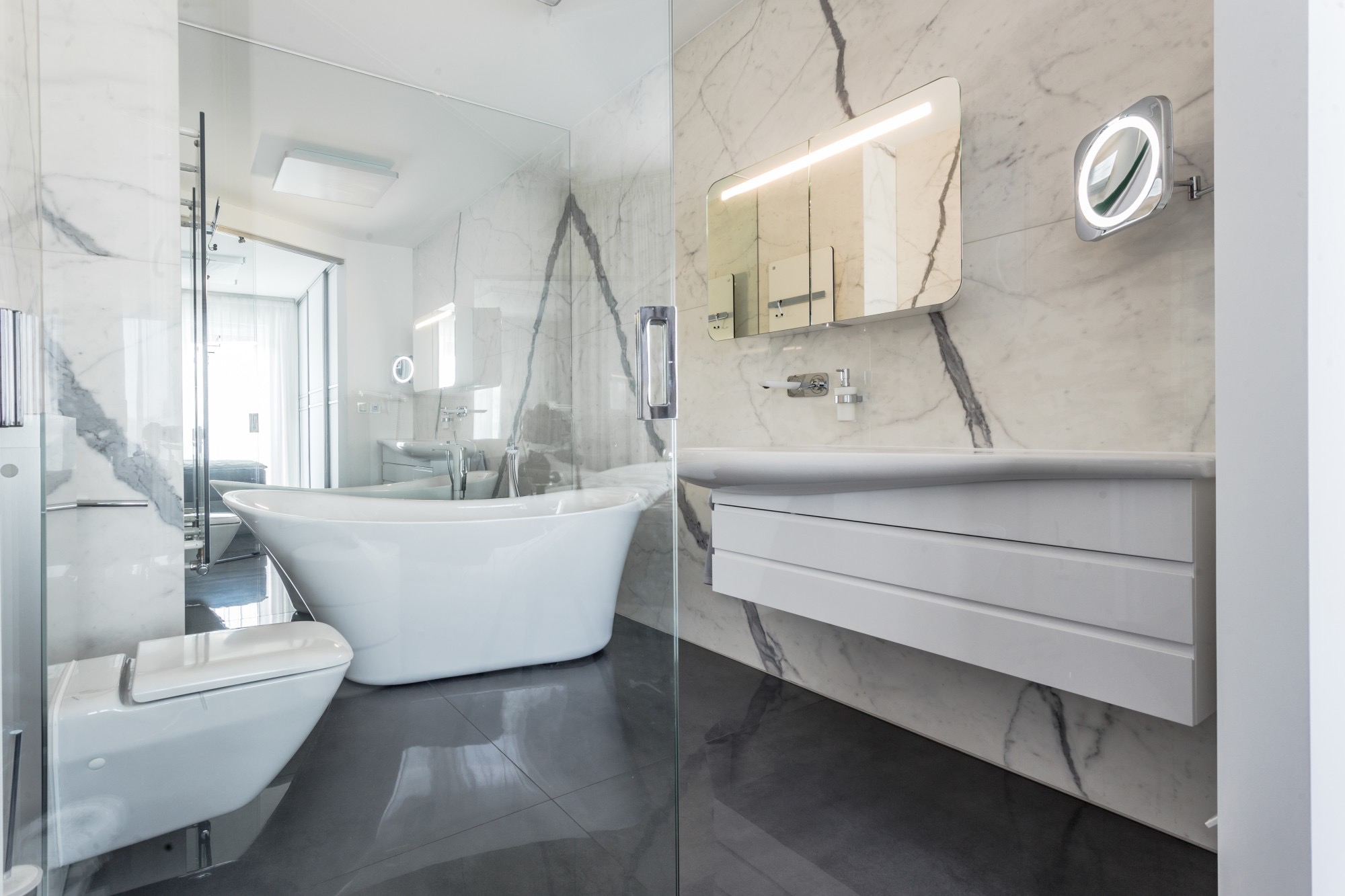 Nowoczesna łazienka z marmurowymi ścianami, wolnostojącą wanną, podwieszaną toaletą i szafką z umywalką, oświetlona lustrami.