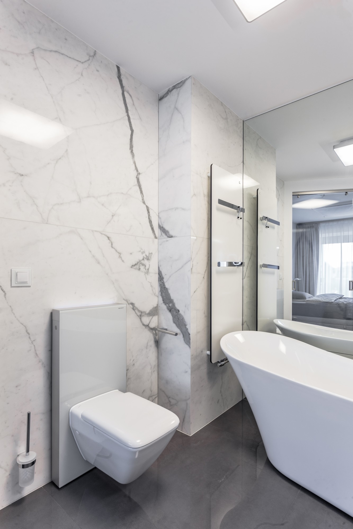 Nowoczesna, minimalistyczna łazienka z białymi marmurowymi ścianami, podwieszaną toaletą, grzejnikiem drabinkowym i wolnostojącą wanną, odbijającą się w lustrze.