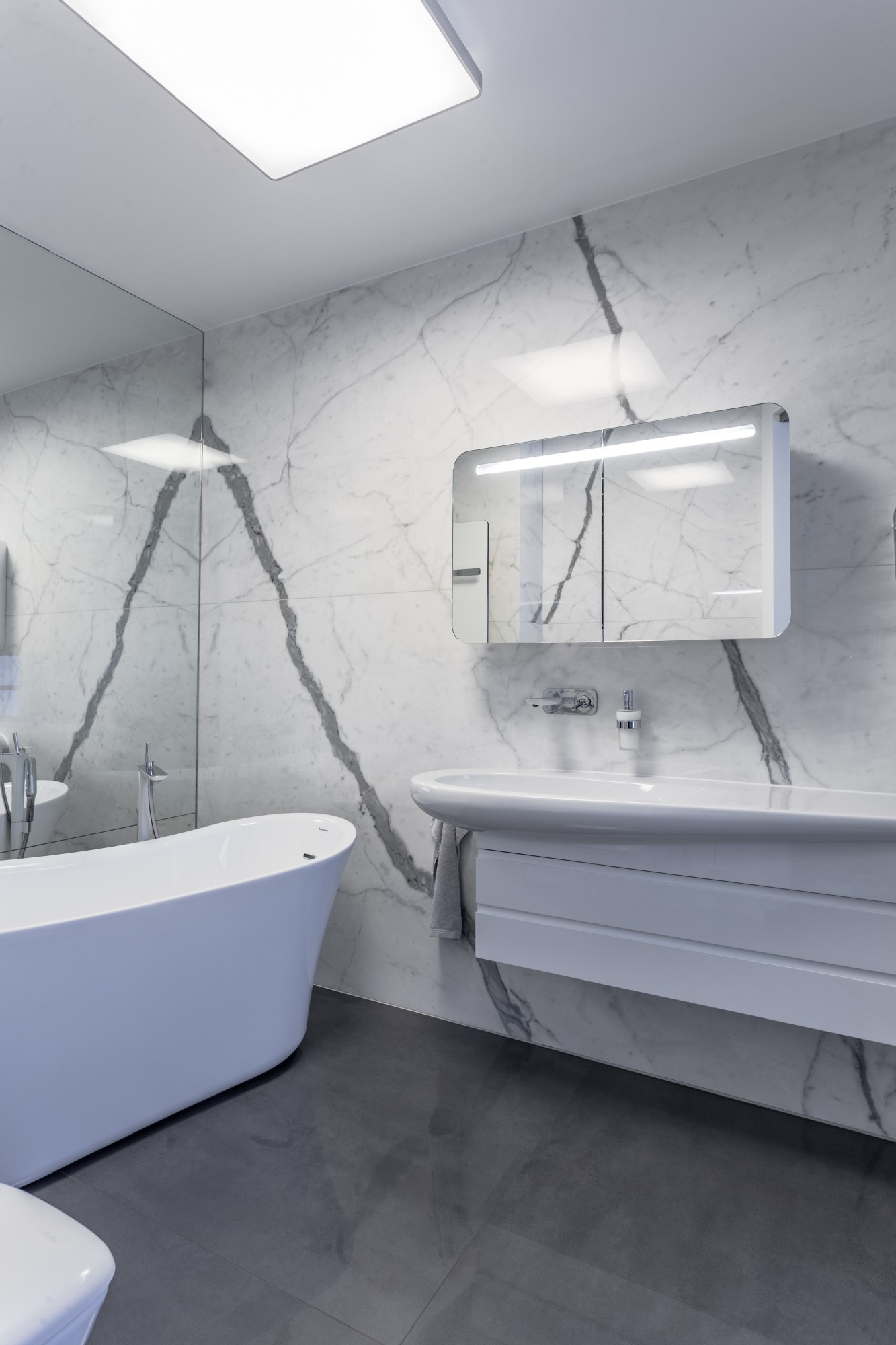 Nowoczesna łazienka z wolnostojącą wanną, umywalką z szafką i lustrem z oświetleniem LED na tle marmurowej ściany z wyraźnym żyłkowaniem.