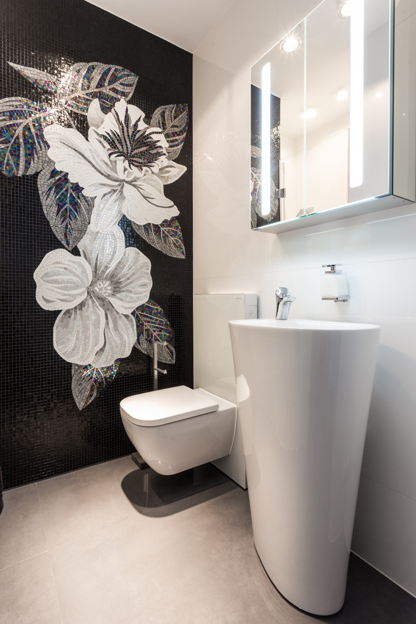 Nowoczesna łazienka z czarną mozaiką przedstawiającą kwiaty, biała umywalka kolumnowa i podwieszana toaleta, oświetlone lustro w szafce.