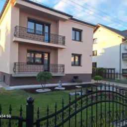 Dwupiętrowy dom z beżową elewacją, ciemnymi oknami i balkonem z ozdobną, kutą balustradą, widoczny zza czarnego, kutego ogrodzenia, z zadbanym trawnikiem i elementami dekoracyjnymi w ogrodzie.