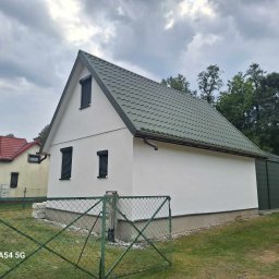 MM MARCIN MAREK - Biały budynek z zielonym dachem i roletami, widziany zza zielonego ogrodzenia z siatki na tle pochmurnego nieba.