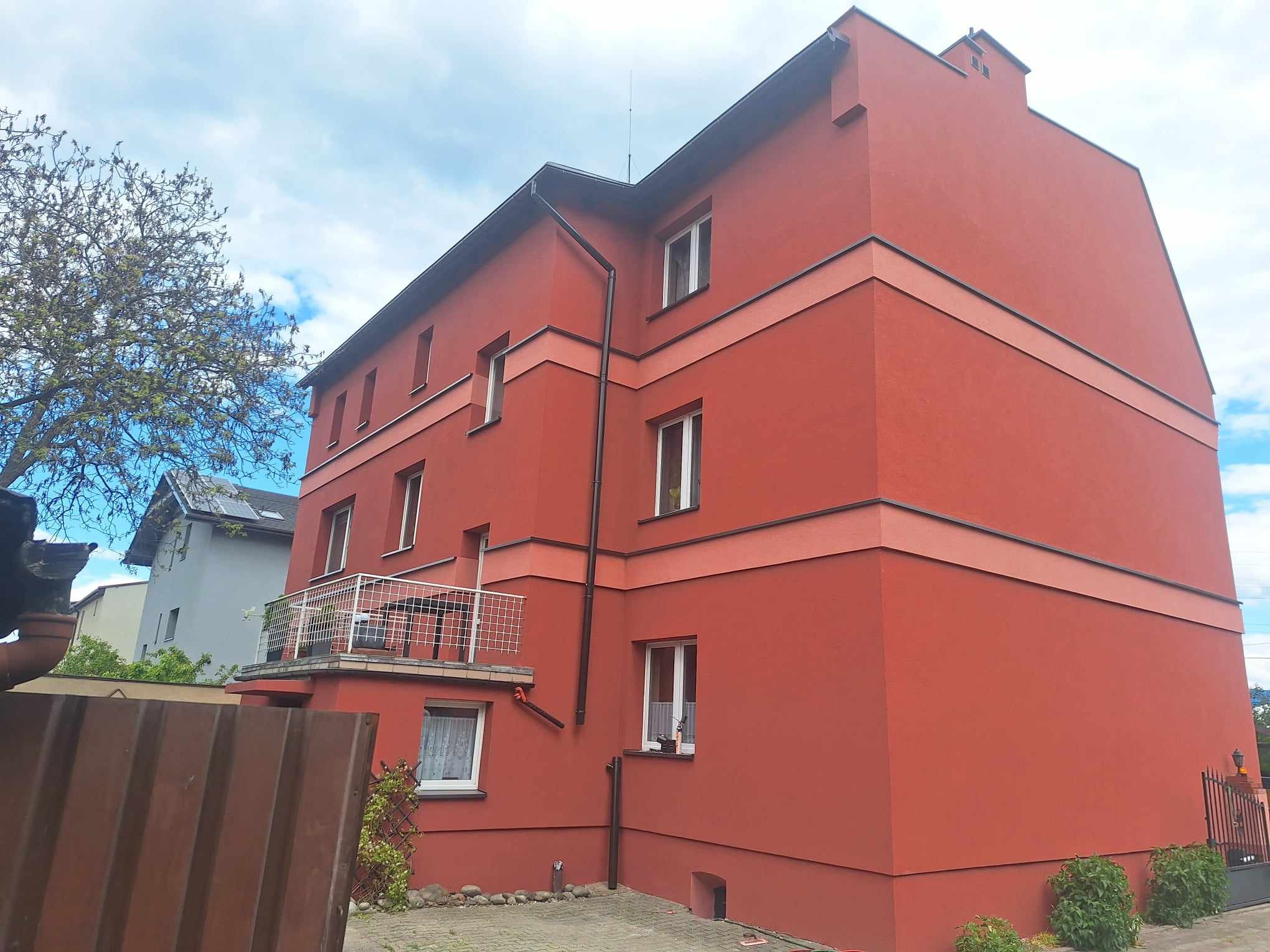 Trzypiętrowy budynek mieszkalny z balkonem, pomalowany na kolor ceglastoczerwony z poziomymi pasami w odcieniu różowym, widok z dołu, z fragmentem ogrodzenia w pierwszym planie.