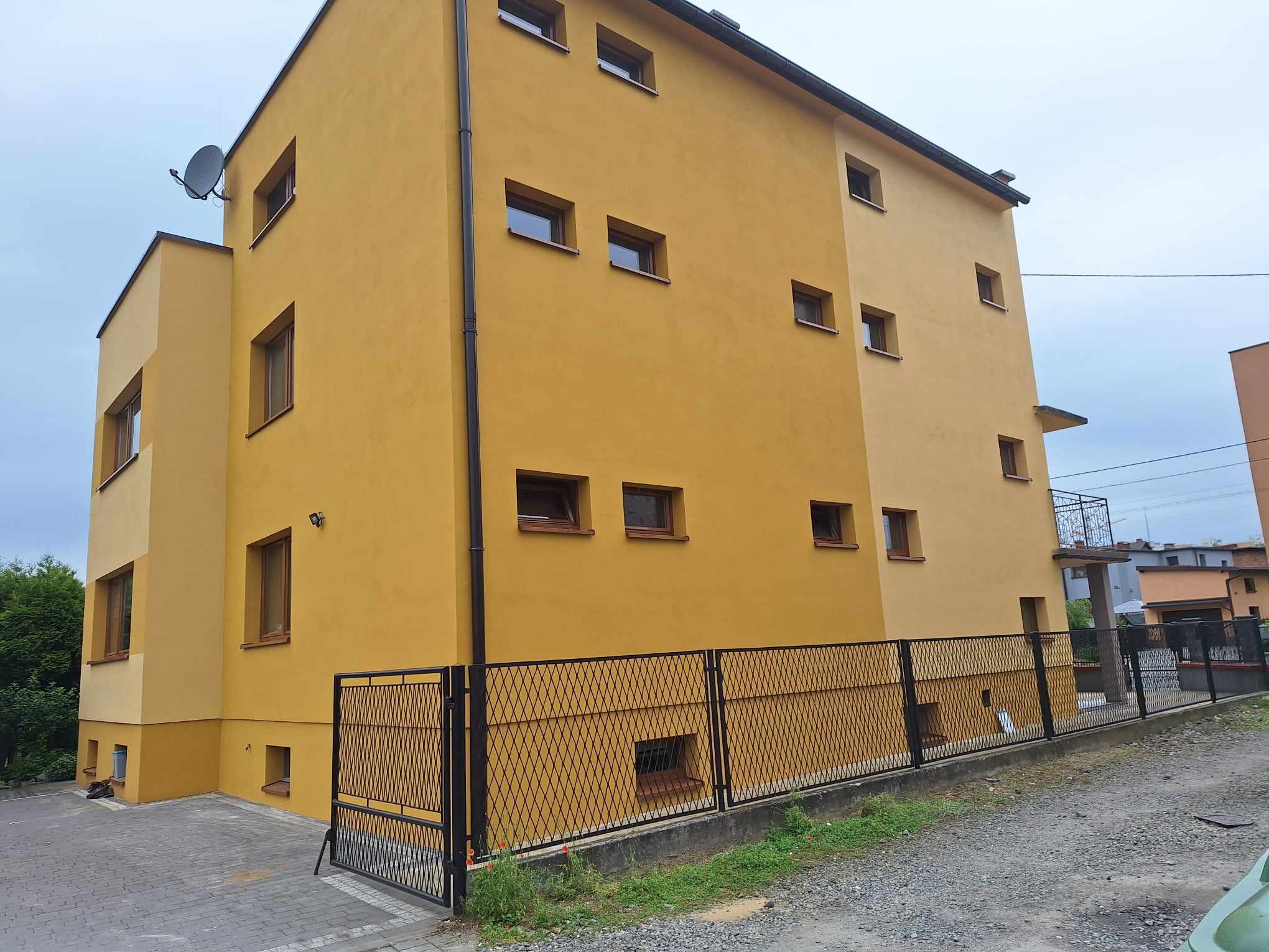 Dwukolorowa elewacja budynku z licznymi oknami, widoczna zza czarnego ogrodzenia z siatki romboidalnej, z anteną satelitarną na dachu.