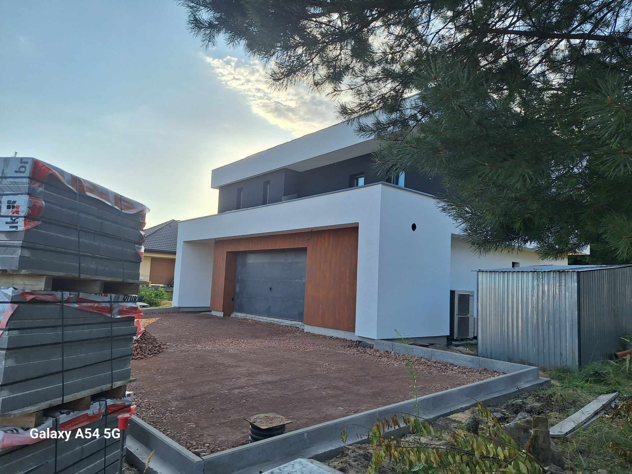 Dwukondygnacyjny dom z płaskim dachem, z garażem z brązową okładziną, widoczne palety z kostką brukową przed domem i przygotowane podłoże pod ułożenie kostki.