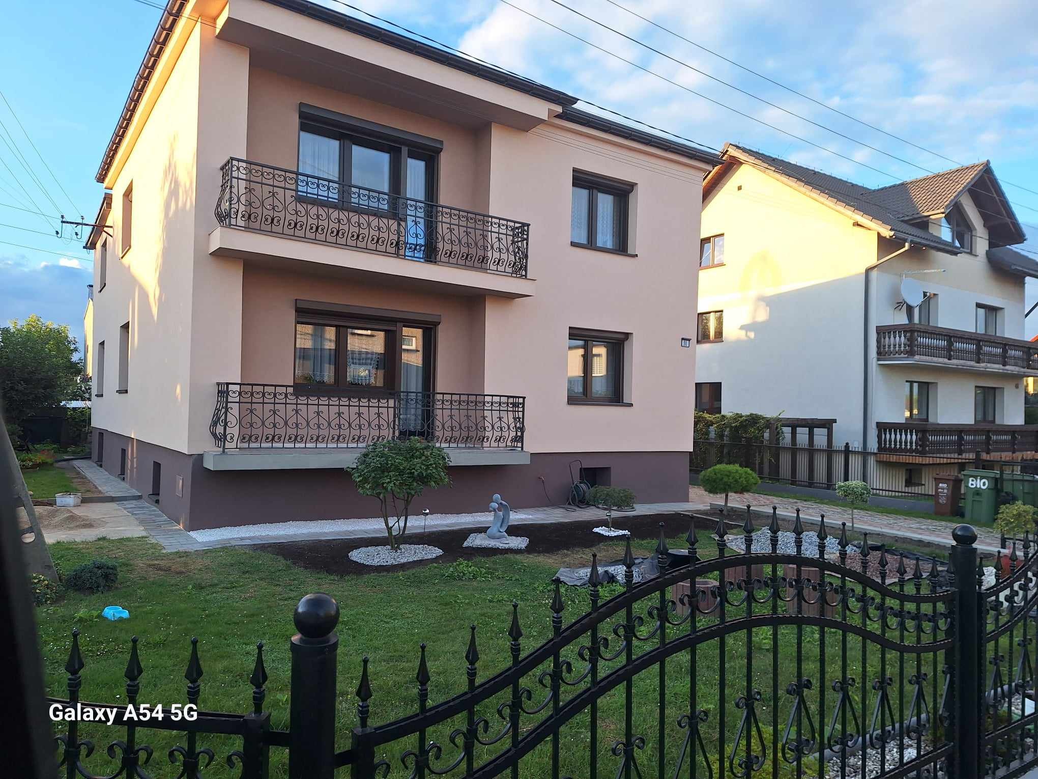 Dwupiętrowy dom z beżową elewacją, ciemnymi oknami i balkonem z ozdobną, kutą balustradą, widoczny zza czarnego, kutego ogrodzenia, z zadbanym trawnikiem i elementami dekoracyjnymi w ogrodzie.