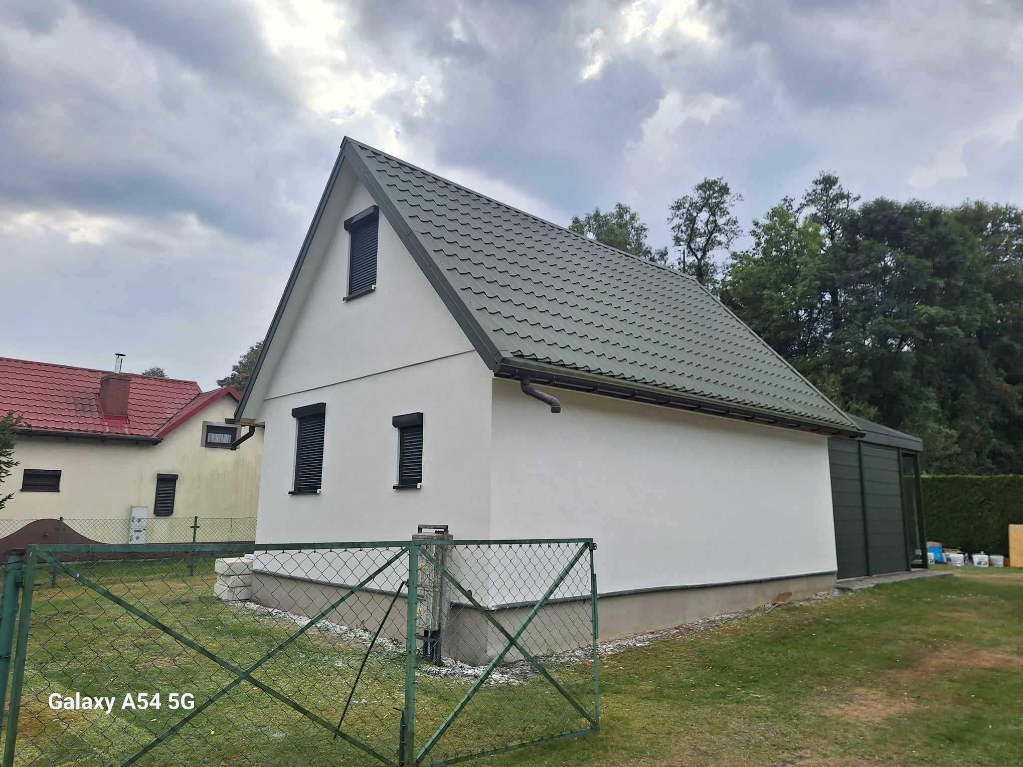 Biały budynek z zielonym dachem i roletami, widziany zza zielonego ogrodzenia z siatki na tle pochmurnego nieba.