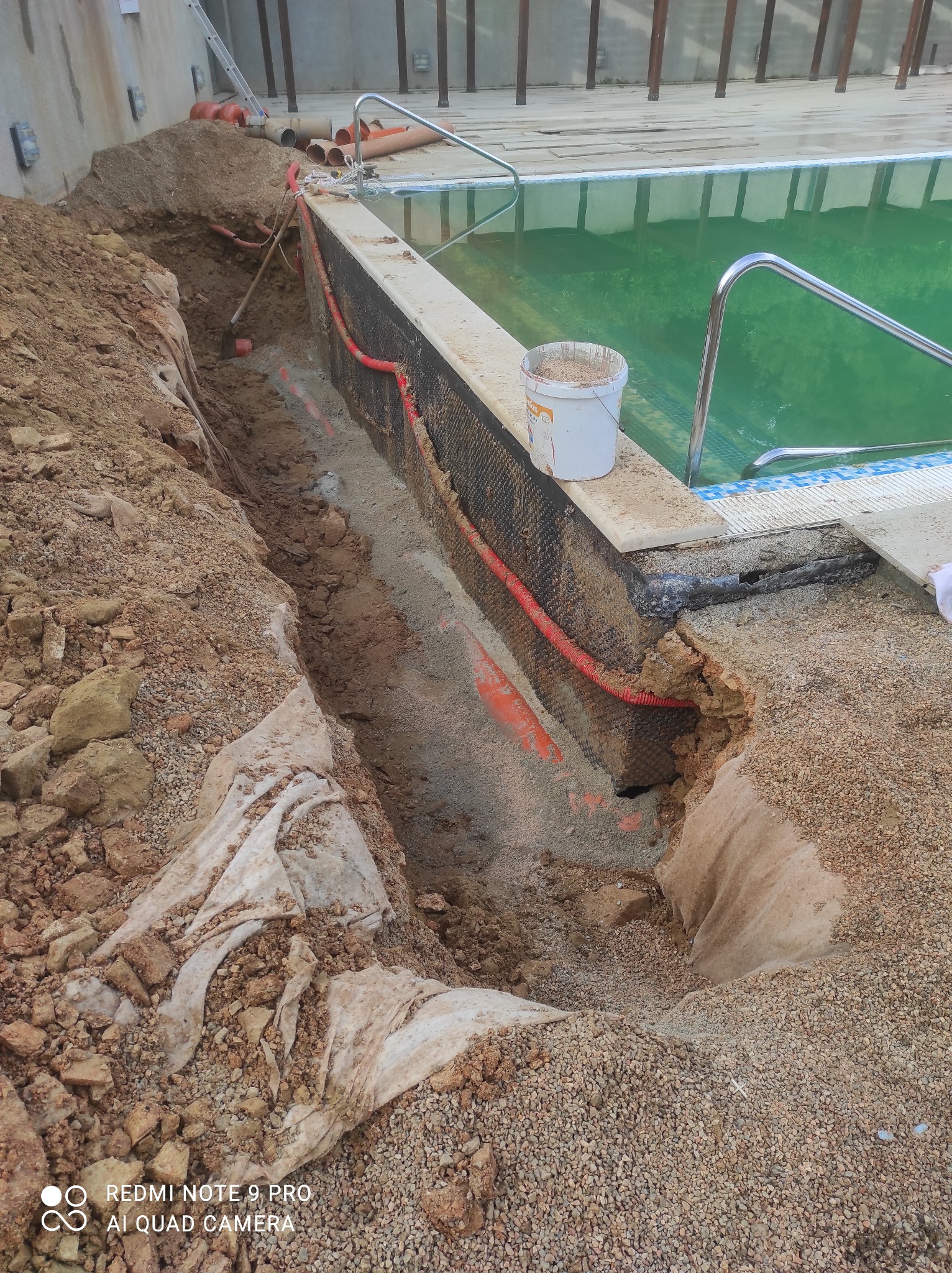 Przygotowanie pod instalację rur w wykopie obok basenu, widoczne rury w pomarańczowej osłonie karbowanej, białe wiadro na krawędzi basenu i ziemia z kamieniami