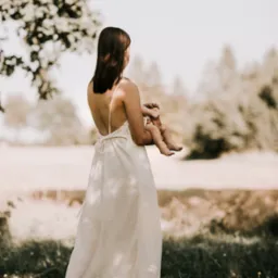 Kobieta w zwiewnej sukience z odkrytymi plecami trzymająca niemowlę na tle rozświetlonej łąki i drzew