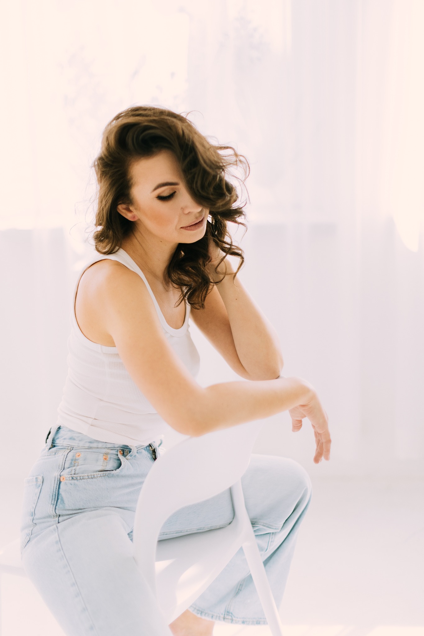 Portret kobiety z brązowymi włosami, siedzącej na białym krześle w jasnym pomieszczeniu, ubrana w biały top i jasne jeansy.